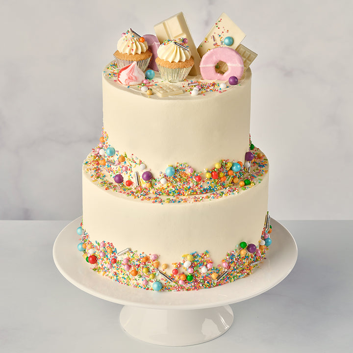 A two-tier vanilla funfetti cake decorated with white chocolate bars, party rings, a mini cupcake, mini meringues, and funfetti sprinkles on top, presented on a white cake stand.