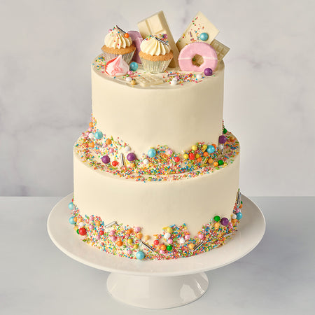 A two-tier vanilla funfetti cake decorated with white chocolate bars, party rings, a mini cupcake, mini meringues, and funfetti sprinkles on top, presented on a white cake stand.