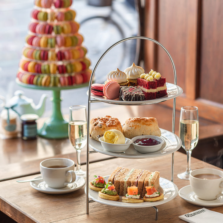 Three-tiered tray with afternoon tea treats including sandwiches, scones, and macarons on a wooden table.