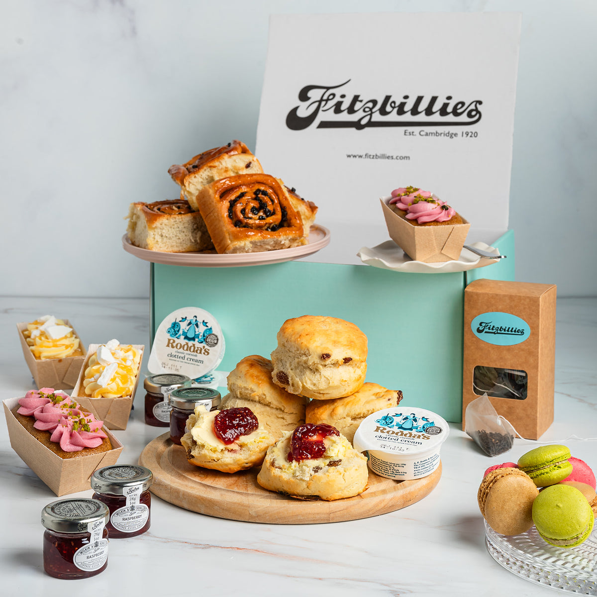 A Fitzbillies bakery display featuring assorted baked goods including scones with jam, Chelsea buns, mini loaf cakes, macarons, and jars of preserves, arranged around a mint green box with a Fitzbillies sign in the background.