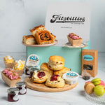 A Fitzbillies bakery display featuring assorted baked goods including scones with jam, Chelsea buns, mini loaf cakes, macarons, and jars of preserves, arranged around a mint green box with a Fitzbillies sign in the background.