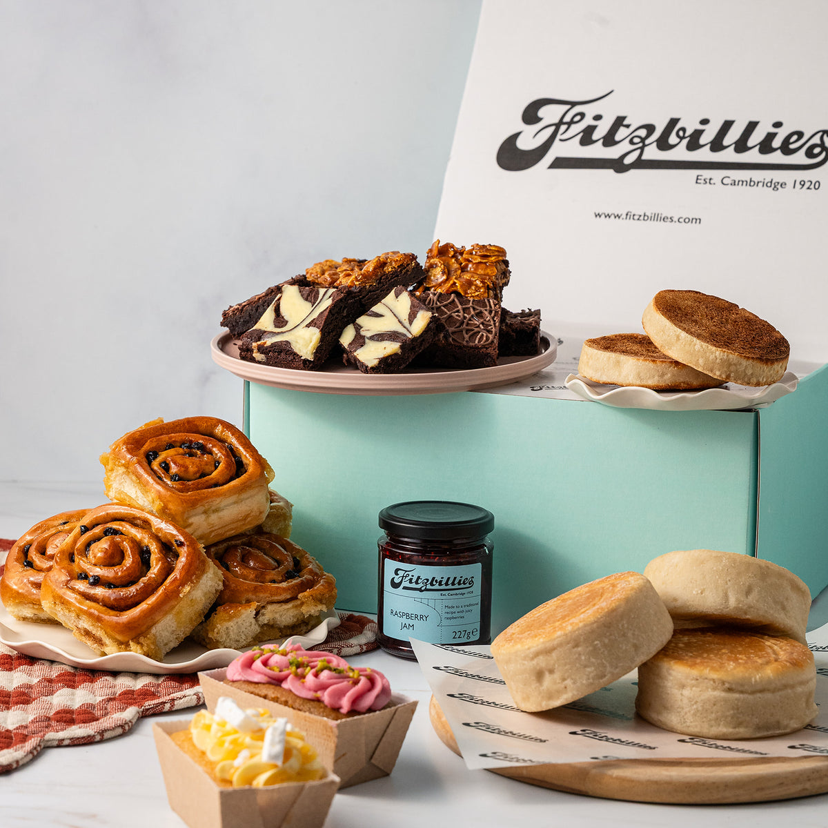 A Fitzbillies bakery gift assortment featuring various baked goods including scones, Chelsea buns, marbled brownies, and mini loaf cakes, arranged around a light blue box and a white branded box on a white background.