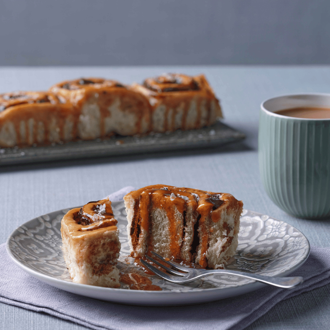 Chelsea bun on a plate with a fork, oozing with syrup, paired with a hot drink