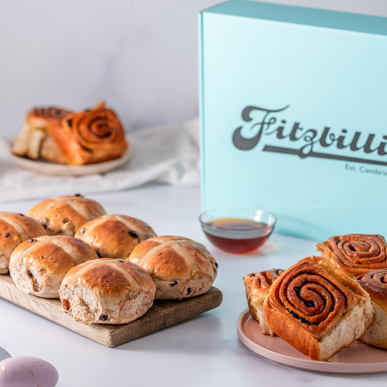 A box of Fitzbillies brand Easter buns, containing hot cross buns and Chelsea buns, presented on a wooden board and plates.