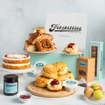 A styled Fitzbillies tea and scone gift assortment on a white marble surface against a pale blue background. Visible items include assorted scones and Chelsea buns on plates and a wooden board, a sponge cake on a cake stand, jars of jam, tea bags, macarons, and branded boxes and cards.