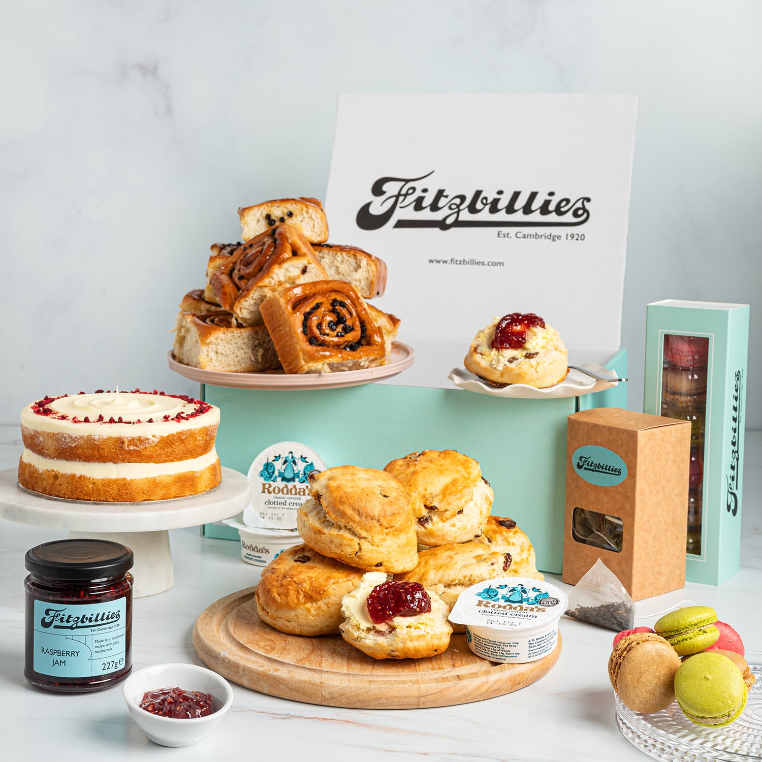 A styled Fitzbillies tea and scone gift assortment on a white marble surface against a pale blue background. Visible items include assorted scones and Chelsea buns on plates and a wooden board, a sponge cake on a cake stand, jars of jam, tea bags, macarons, and branded boxes and cards.
