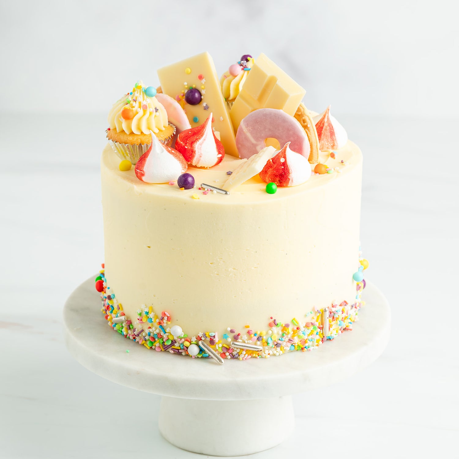 Decorative cake with colourful sprinkles, meringues, mini cupcakes and party rings on a white background