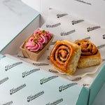 Two Chelsea Buns and a pink frosted mini loaf cake displayed inside an open Fitzbillies box lined with branded paper. The products include a Chelsea bun with currants and a pink pistachio and raspberry cake topped with nuts.