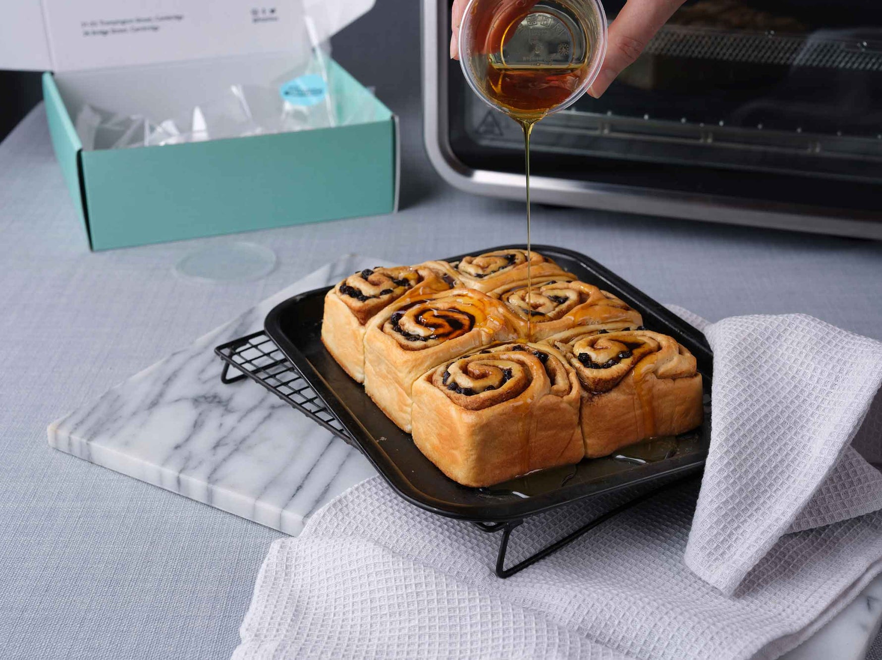 Pouring syrup over bake at home Chelsea buns cooling on an oven tray rack