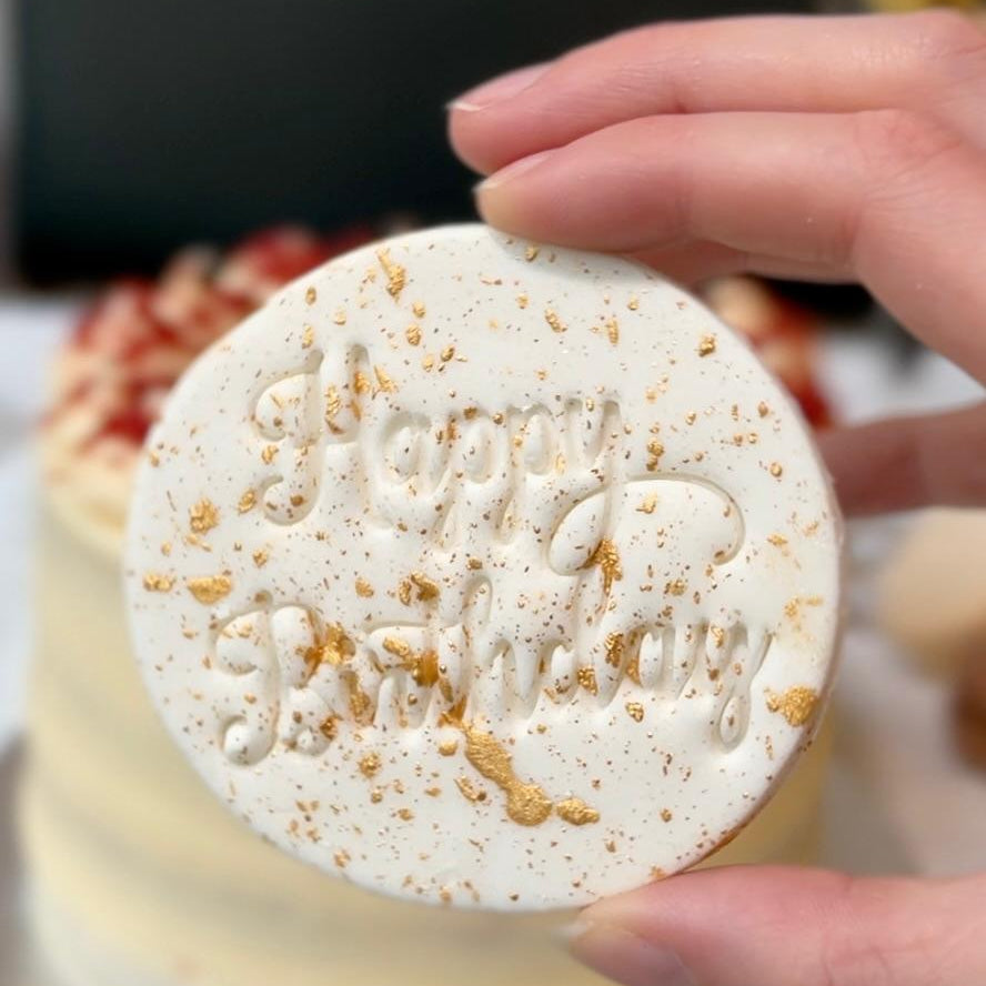 Hand holding a round, white chocolate plaque with gold specks and 'Happy Birthday' text against a blurred background.