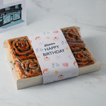 A gift box of cinnamon rolls wrapped in clear plastic with a white paper band that says 'Happy Birthday' and has small birthday-themed illustrations. A small card is partially visible in the background on a light marble surface.