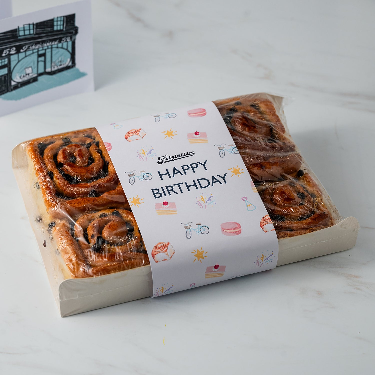 A gift box of cinnamon rolls wrapped in clear plastic with a white paper band that says 'Happy Birthday' and has small birthday-themed illustrations. A small card is partially visible in the background on a light marble surface.