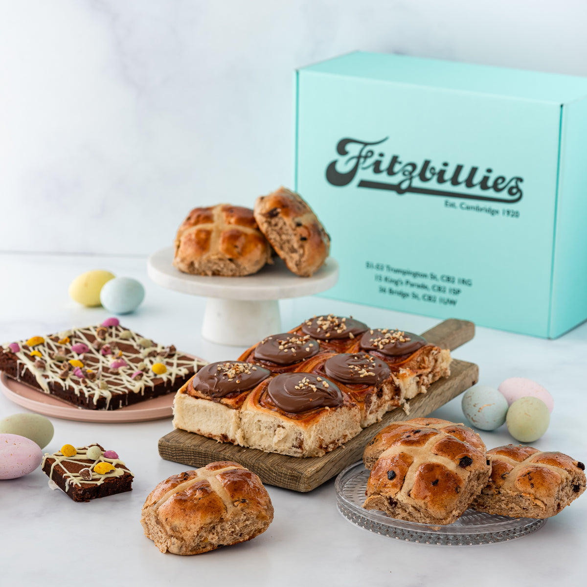 An assortment of Easter chocolate treats including hot cross buns, Nutella buns, and chocolate brownies, presented in front of a blue gift box with the 'Fitzbillies' logo.